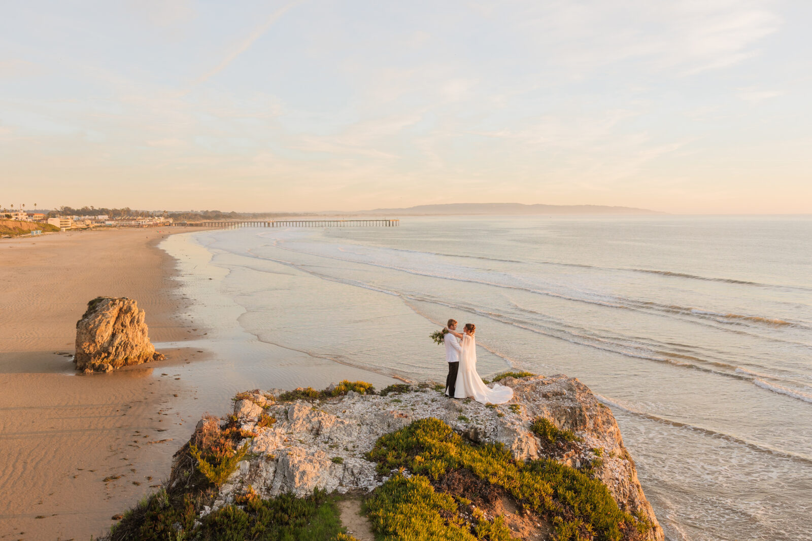 Bride and groom standing together on a scenic beach cliff overlooking the ocean as sunset, with soft golden light, flowing dress, and waves crashing below, capturing the romantic and natural wedding moment