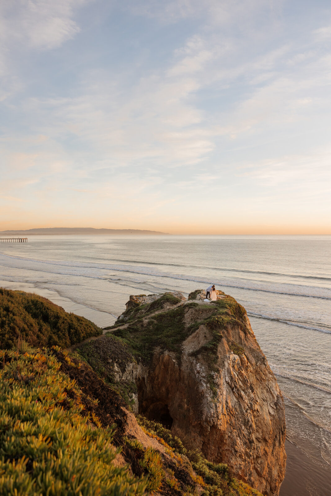 Bride and groom standing on cliffside overlooking the ocean at sunset in Pismo Beach, California, romantic moment captured by Southern California wedding photographer